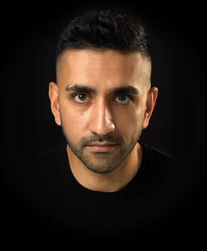 Susheel Kumar - Scottish and Indian English Voiceover Artist - headshot, close front-facing, looking into the camera wearing a black top that blends into a black background.